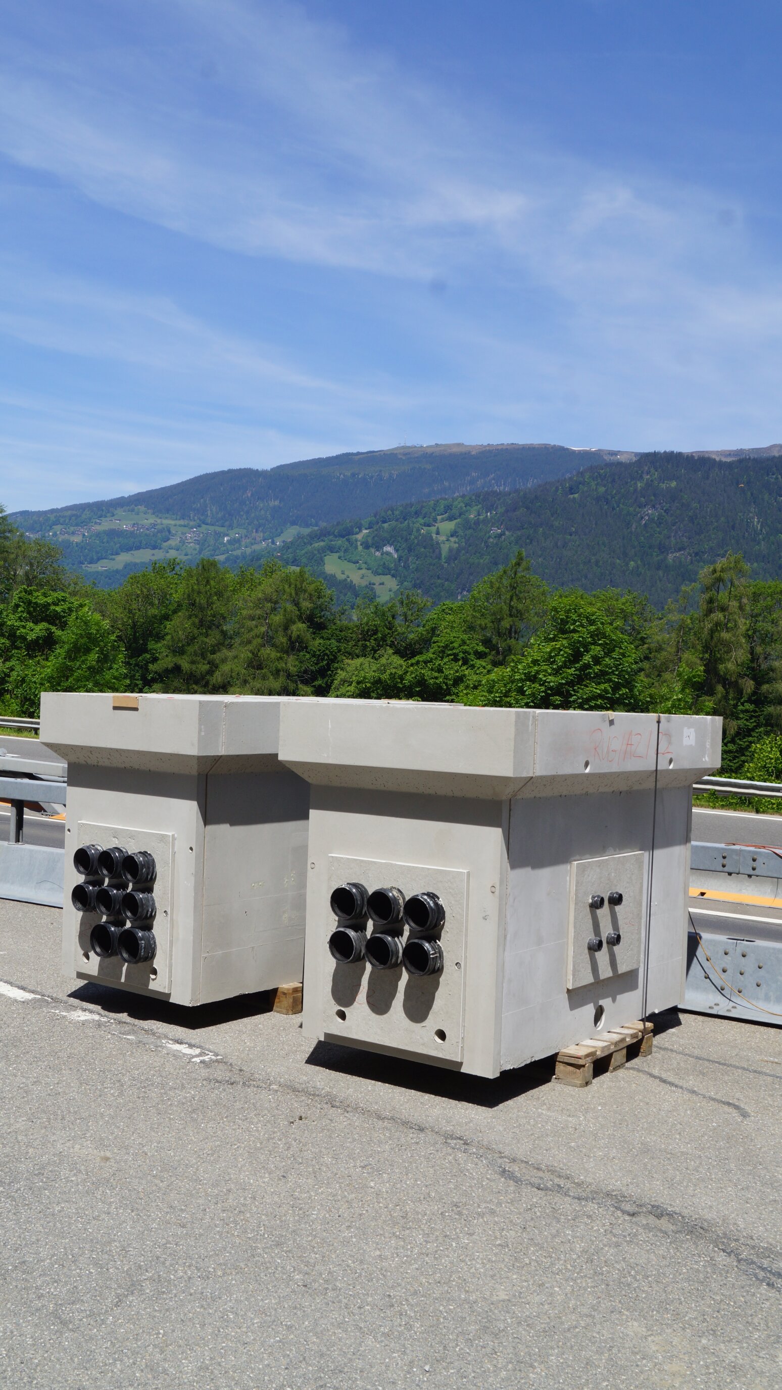 Zwei große Betonkästen mit mehreren schwarzen Rohranschlüssen an den Seiten stehen auf Paletten in der Nähe einer Straße, mit grünen Bäumen und Bergen im Hintergrund unter einem blauen Himmel.